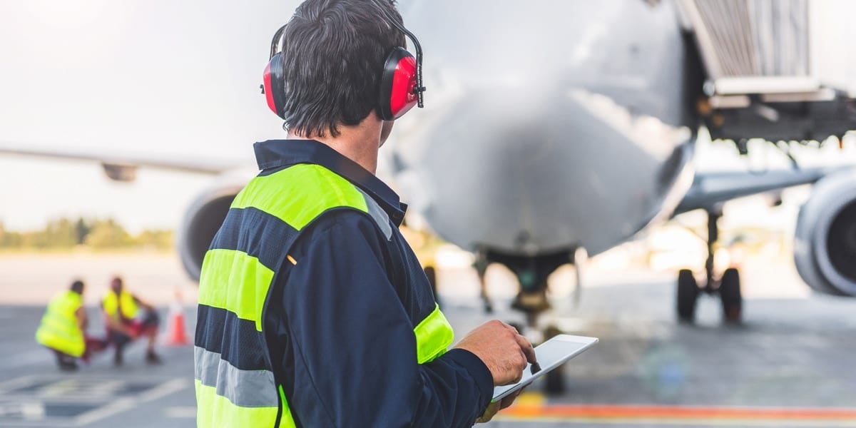 man with ear protection and high vis observing men working on aeroplane